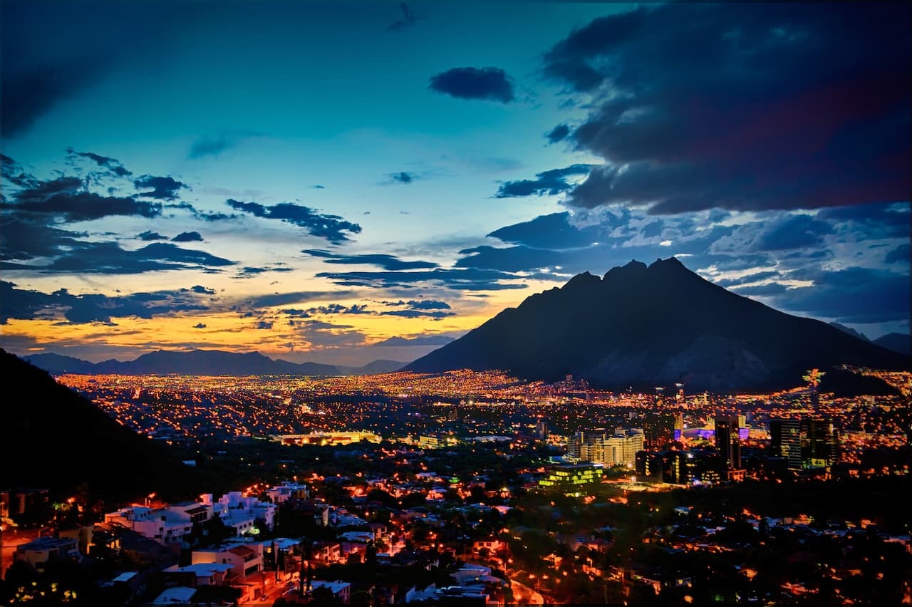 Panorama de la ciudad de Monterrey al atardecer, con montañas de fondo y luces brillando en la ciudad.