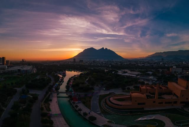 Vista panorámica de la ciudad de Monterrey al atardecer, con el Cerro de la Silla y un río que atraviesa un área verde y construcciones en primer plano.