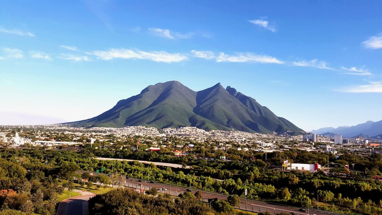 Paisaje que muestra montañas verdes y una ciudad en el primer plano, con un cielo azul despejado y algunas nubes dispersas.