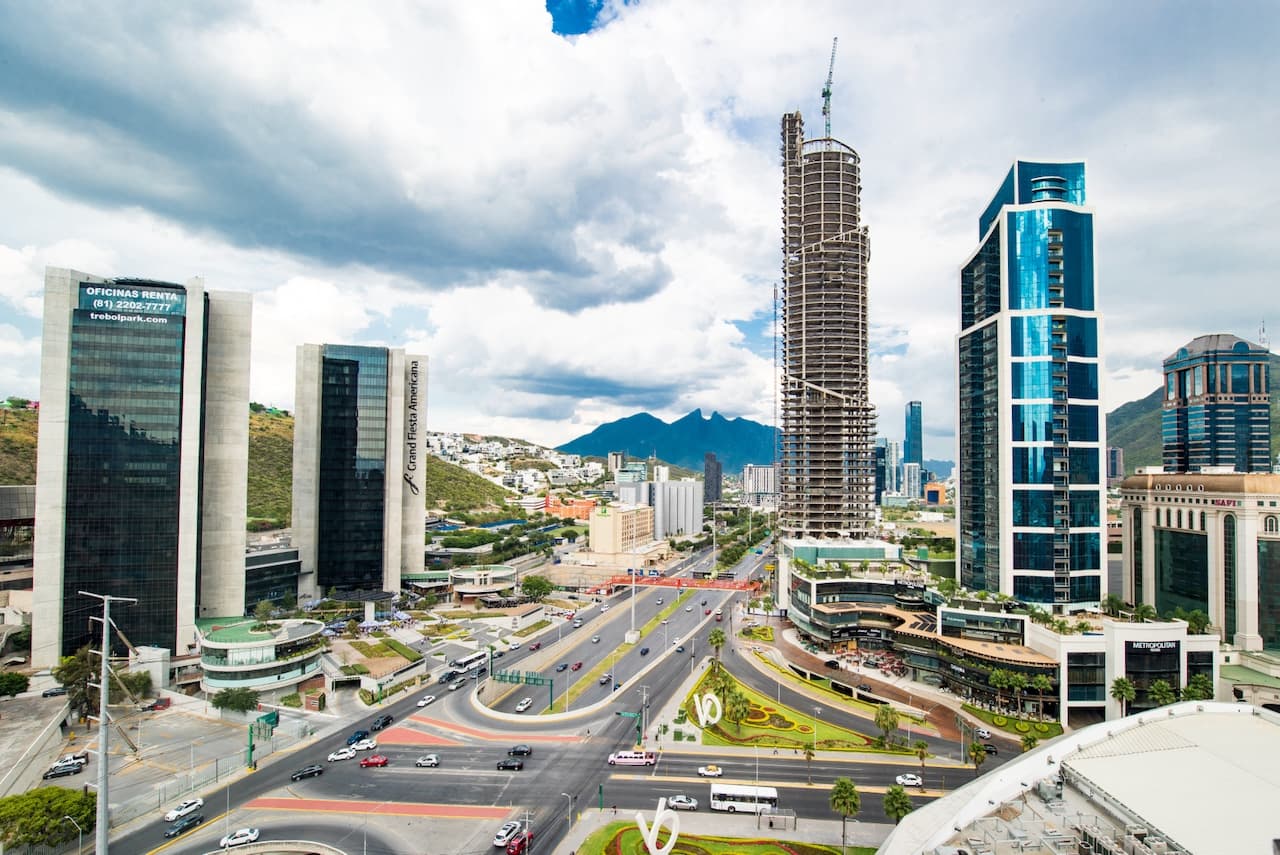 Vista panorámica de una zona urbana en Monterrey, México, con edificios modernos, tráfico vehicular y montañas al fondo. Se destaca una construcción en proceso de un rascacielos.