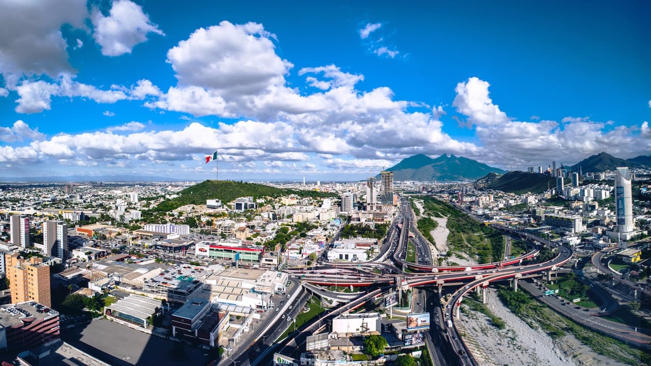 Vista panorámica de Monterrey, México, con edificios, montañas y una bandera mexicana ondeando. Se observan carreteras y puentes en el paisaje urbano.